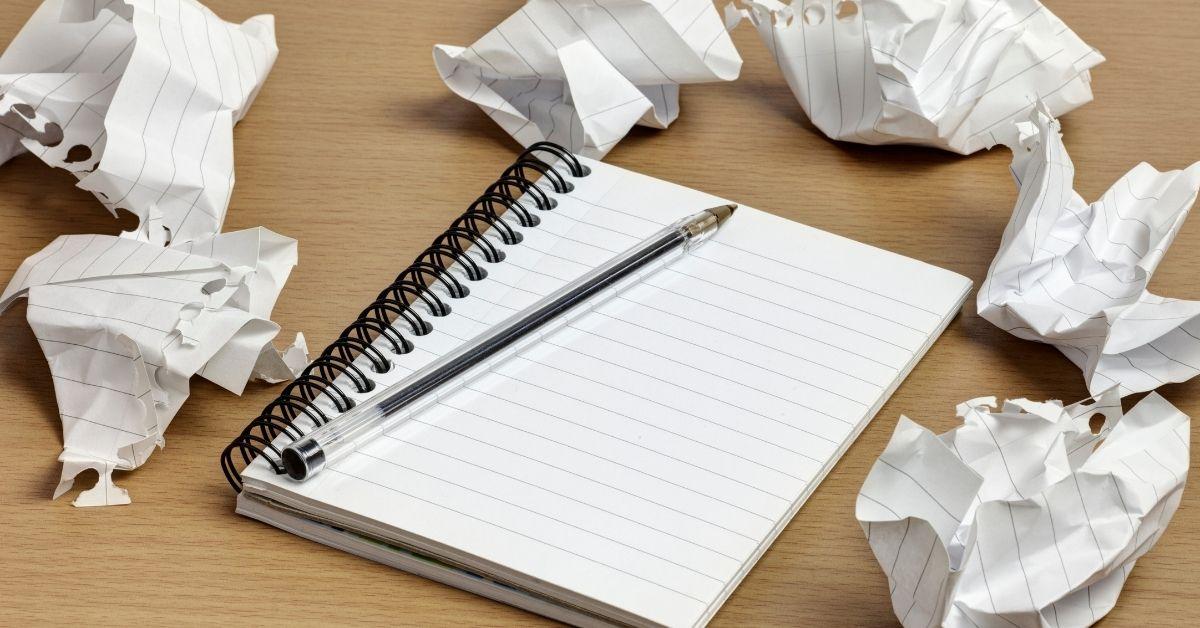 A lined notebook on a table with a pen resting on it. The notebook is surrounded by crumpled pieces of paper containing blog drafts.